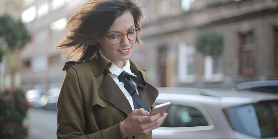 woman with short brown hair wearing glasses and coat using her smartphone while walking down the street